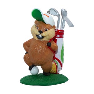 May include: A brown beaver figurine wearing a white and green golf cap, standing on a green base with one foot on a white golf ball. Beside the beaver is a white, red, and green golf bag containing two silver-colored golf clubs with visible detail.
