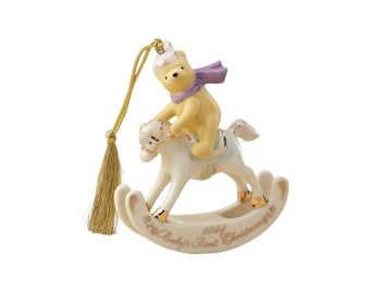 May include: A ceramic Christmas ornament featuring a yellow bear wearing a white hat and purple scarf, riding a white rocking horse. The base of the rocking horse is inscribed with the text 2024 Baby's First Christmas. A gold tassel hangs from the top.