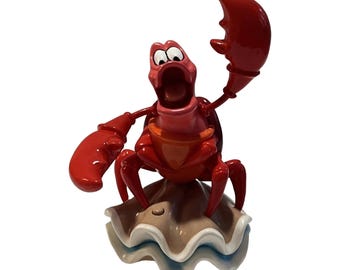 Peut inclure: Une figurine de crabe rouge brillant avec de grandes pinces, de grands yeux et la bouche ouverte, posée sur une base en coquillage festonné beige et blanc. Le crabe a une finition lisse et brillante, une pince levée en l'air.
