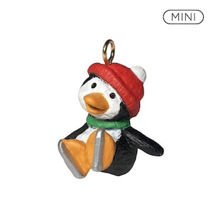 Può includere: Una piccola statuetta di pinguino con un berretto di lana rosso con pompon bianco, una sciarpa verde e piedi arancioni con pattini da ghiaccio argentati. Il pinguino ha il corpo bianco e nero e il becco arancione. La parola MINI è stampata in nero.