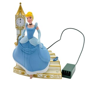 May include: A figurine of Cinderella wearing a light blue gown and gloves, standing on a staircase base next to a tall clock tower. A single blue glass slipper rests on the stairs. A black power cord extends from the base to a green battery pack.