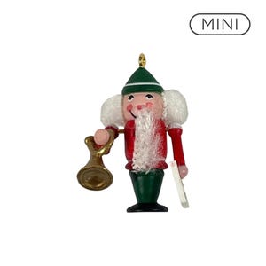 May include: A miniature nutcracker ornament featuring a green hat, red jacket, and green trousers. The figure has white hair and a beard, holding a gold-colored bell in one hand and a small white rectangular object in the other. The text MINI is visible.