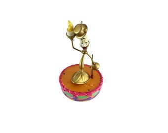 Puede incluir: Una figura de candelabro antropomórfico de color dorado con rostro sonriente, que lleva un pequeño sombrero de copa y sostiene una llama amarilla. La figura se asienta sobre una base redonda naranja decorada con un borde festoneado rosa, verde y morado.
