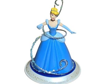 Peut inclure: Une figurine représentant un personnage aux cheveux blonds relevés, portant une robe bleue et des gants assortis. Un tourbillon translucide et scintillant entoure la silhouette. La base est circulaire, blanche, avec le texte A princess in the making.