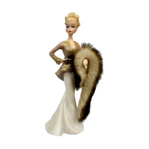 May include: A figurine of a blonde person with an updo hairstyle wearing a tan jacket with gold embroidery and a long white skirt. The figure holds a long, textured brown and cream-colored faux fur stole draped over one arm, standing upright.