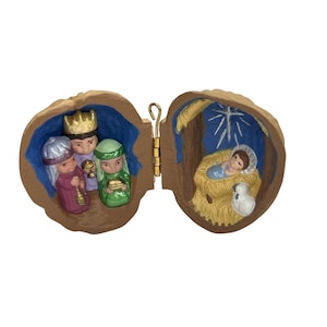 May include: A miniature Christmas nativity scene inside a walnut shell. The left side features three figures in colorful robes, while the right side depicts baby Jesus in a manger with a lamb. The shell is hinged with a gold-colored clasp.