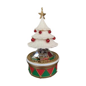 May include: A decorative tabletop Christmas tree ornament featuring a cream-colored textured tree topped with a gold star and adorned with small red spherical ornaments. The base is a red and green drum with a miniature train circling the center.