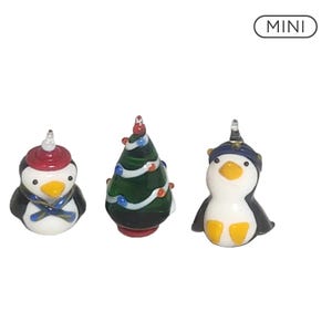 May include: Three glass figurines labeled MINI, including two penguins and one Christmas tree. One penguin wears a red hat and blue scarf, the other a blue hat. The green tree features white garland and small red and blue ornaments.