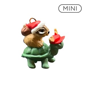 Peut inclure: Une figurine miniature représentant un tamia brun portant un bonnet de Père Noël rouge et blanc, chevauchant une tortue verte coiffée d'une casquette rouge à petites cornes. Le mot MINI est inscrit dans un ovale noir en haut de l'image.