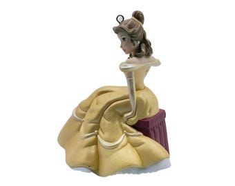 Puede incluir: Una figura de un personaje con cabello castaño recogido en un moño, vistiendo un vestido amarillo de hombros descubiertos con ribetes blancos. El personaje está sentado sobre una base rectangular de color rojo oscuro. Un pequeño aro metálico está sujeto a la parte superior de la cabeza.