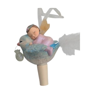 May include: A decorative bottle stopper featuring a light blue bird with glittery accents, carrying a small white pouch in its beak. A small figure in a light pink outfit rests on the bird's back, with white ribbon and feather details attached.