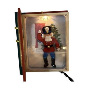 May include: A light-up decorative book-shaped display featuring a nutcracker figure in a red uniform with blue pants and black boots. The interior scene shows a Christmas tree, pink curtains, and a lit chandelier, all encased in a frame.