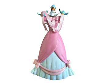 Puede incluir: Una figura decorativa que presenta un vestido rosa y azul claro con falda drapeada y lazos. Un pequeño personaje con sombrero azul se sienta en la parte superior, flanqueado por dos pájaros azul claro. Una cinta rosa cuelga de los hombros.