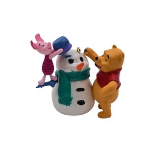 May include: A decorative figurine featuring a yellow bear in a red shirt and a small pink pig character. They are standing on either side of a white snowman with a green scarf, an orange carrot nose, and black coal buttons. The pig holds a blue hat.