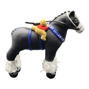 Könnte beinhalten: Eine schwarze Pferdefigur mit weißer Schnauze und weißem Behang an den Hufen. Ein kleiner gelber Bär in einem roten Hemd sitzt auf dem Rücken. Das Pferd trägt ein blaues Geschirr mit der Aufschrift 2014 und silbernen Akzenten sowie einen schwarzen Zaum.