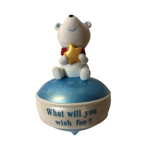 May include: A white polar bear figurine with a red scarf and a gold star sits atop a blue, rounded base decorated with white snowflakes. The base features the text "What will you wish for?" printed in blue lettering against a white background.