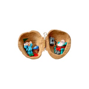 May include: A miniature Christmas-themed ornament shaped like a walnut shell. Inside, a figure in blue pajamas is on the phone, and Santa Claus is seated in a chair. The ornament is hinged with a gold-colored clasp.