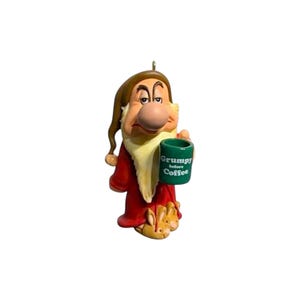 May include: A figurine of a character with a long white beard, wearing a brown nightcap and a red robe, holding a green mug with the text Grumpy before Coffee printed in white. The character wears tan slippers shaped like rabbit faces on their feet.