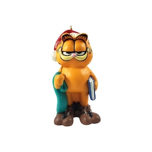 May include: A small orange Garfield figurine wearing a red Santa hat with a white trim. The character holds a blue book in one hand and a green fabric item in the other, while wearing large brown shoes. A gold loop is attached to the top of the hat.