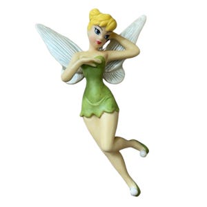 May include: A figurine of a fairy with blonde hair styled in a bun, wearing a green dress with a jagged hemline. The figure has large, translucent white wings, light skin, and small green shoes with white pom-poms at the tips of the toes.