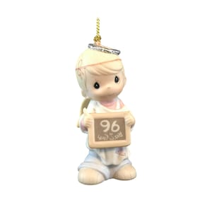 May include: A porcelain figurine of a child angel with blonde hair, wearing a silver halo and holding a rectangular sign with the number 96 and the text Peace on Earth. The ornament features a gold hanging cord attached to the top of the halo.