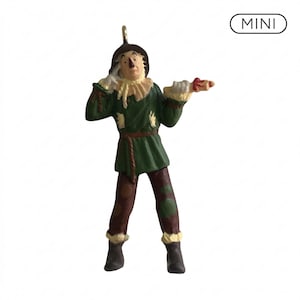 Puede incluir: Una figura miniatura de espantapájaros que viste una túnica verde oscuro, pantalones marrones con parches y un sombrero marrón. La figura tiene un cuello de paja, botas oscuras y sostiene un pequeño pergamino. La palabra MINI aparece arriba a la derecha.