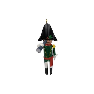 May include: A Christmas ornament shaped like a toy soldier. The soldier wears a black hat, green jacket with gold trim, red sash, and white pants. He holds a blue telescope to his eye. A gold loop is at the top for hanging.