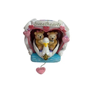 Puede incluir: Una figura decorativa que muestra dos ositos de peluche de color marrón claro sentados dentro de un bote con forma de cisne blanco. La estructura está enmarcada por un borde en forma de corazón con el texto Sweethearts y un dije de corazón rosa.