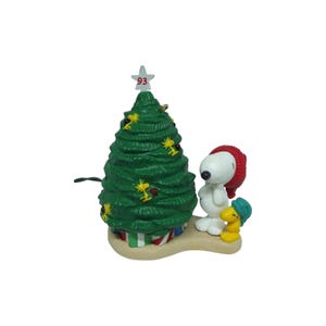 May include: A Snoopy and Woodstock Christmas tree figurine featuring a green tree decorated with small yellow Woodstock figures, red ornaments, and a white star topper with the number 93. Snoopy wears a red hat, and Woodstock wears a green hat.