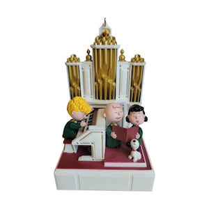 May include: A Peanuts character figurine featuring Schroeder playing a white and gold pipe organ while Charlie Brown and Lucy sing from a red book, accompanied by Snoopy. The scene is set on a red and white base with a metallic hanging loop.