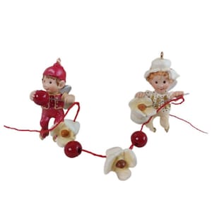May include: Two small elf-like figurines connected by a red string featuring white flower-shaped beads and red spherical beads. The left figurine wears a red outfit and hat, while the right figurine wears a white outfit and hat with gold accents.