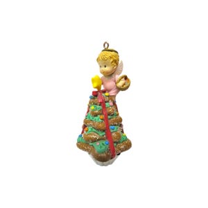 Könnte beinhalten: Ein hängendes Ornament mit einem blonden Engel mit weißen Flügeln in einem rosa Kleid, der einen kleinen Korb hält. Der Engel steht auf einem braunen, kegelförmigen Baum, der mit grünem Zuckerguss, bunten Streuseln, einem roten Band und einem gelben Stern verziert ist.