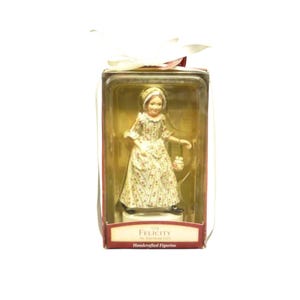 Pode incluir: Uma estatueta artesanal Felicity American Girl numa caixa de plástico transparente com uma fita branca no topo. A estatueta usa um vestido comprido de cor creme com um pequeno padrão floral vermelho, um gorro a condizer e uma pequena mala.