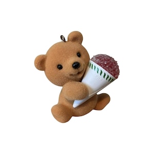 Puede incluir: Un adorno de oso de peluche marrón que sostiene un cono de nieve rojo. El oso tiene una cara sonriente con ojos negros y una nariz negra. El cono de nieve está en un cono de papel blanco con rayas verdes.