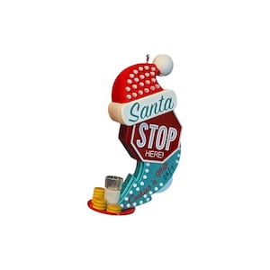 以下が含まれることがあります： サンタの赤い帽子が赤い一時停止標識の上に乗ったクリスマスオーナメント。標識には「Santa STOP HERE!」と書かれ、ティール色の土台には「Cookies n' Milk 24/7」の文字があり、小さなミルクグラスと黄色いクッキーが添えられています。