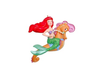 Peut inclure: Une figurine décorative représentant une sirène aux cheveux roux avec une queue verte scintillante chevauchant un hippocampe orange. La sirène porte un haut en forme de coquillage violet et tient un collier de perles blanches.