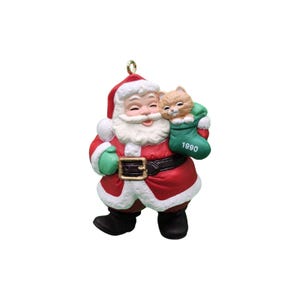 May include: A Santa Claus Christmas ornament wearing a red suit with white trim, a black belt with a gold buckle, and green mittens. Santa holds a green stocking containing a small orange cat, with the year 1990 printed in white on the stocking.