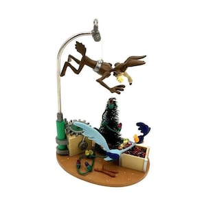 Puede incluir: Una figura decorativa que muestra a un coyote marrón suspendido de un brazo de metal plateado sobre un pequeño árbol de Navidad verde y un pájaro Correcaminos azul. La escena incluye cajas de madera, luces navideñas y una base color canela.