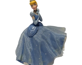 Peut inclure: Une figurine de Cendrillon aux cheveux blonds relevés, portant une robe bleue avec une surcouche transparente et scintillante. Le personnage porte un ras-de-cou noir, des gants longs bleus et un diadème argenté. La robe a une jupe ample.