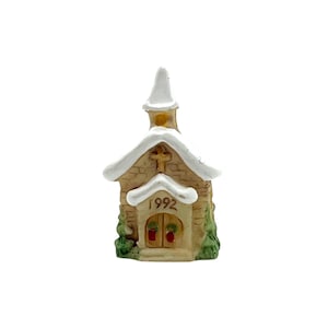 May include: A miniature ceramic church figurine featuring a tan brick facade, a white snow-dusted roof, and a steeple. The front displays a cross, the year 1992, and arched double doors adorned with small green wreaths with red accents on each side.