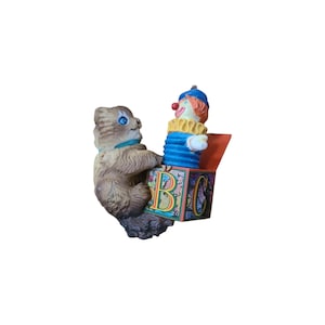 May include: A vintage, whimsical figurine featuring a brown tabby cat and a clown emerging from a colorful ABC block. The cat, with blue eyes and a teal collar, appears to be interacting with the clown. The clown has a blue hat and a ruffled collar.