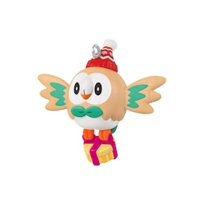 May include: A round, tan bird-like ornament with green wing accents, a white face, and orange feet. It wears a red and white winter hat with a snowflake pattern and holds a yellow gift box tied with a bright pink ribbon, featuring a metal loop.
