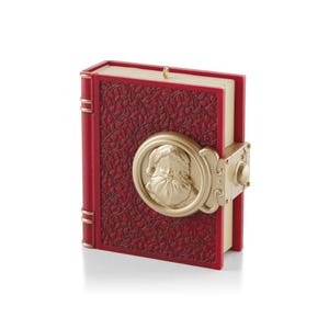 May include: A rectangular red book-shaped ornament featuring a textured cover with an intricate embossed floral pattern. A circular gold-toned medallion depicting a bearded figure in a hat is centred on the front, secured by a gold-toned latch.