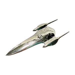 Può includere: Un modello di caccia stellare Naboo N-1 con finitura cromata, punta affusolata, due motori montati lateralmente e una cupola centrale per la cabina di pilotaggio. La superficie è riflettente e mostra linee di pannello e testo vicino al cockpit.