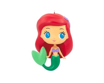 Peut inclure: Une figurine de sirène stylisée avec des cheveux rouges vifs, de grands yeux bleus et un haut en forme de coquillages violets. Le personnage possède une queue vert vif avec une nageoire assortie et un petit accessoire violet dans les cheveux.