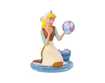 Peut inclure: Une figurine représentant un personnage blond vêtu d'un corsage marron, de manches vert clair et d'un tablier crème, agenouillé sur une base bleue. Le personnage tient une sphère transparente contenant deux danseurs, avec un seau.