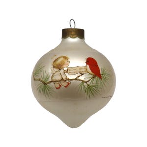 May include: A pear-shaped glass Christmas ornament with a metallic silver finish. The surface features a painted illustration of a small angel and a bright red bird perched on a pine branch, both looking at an open book held by the angel.
