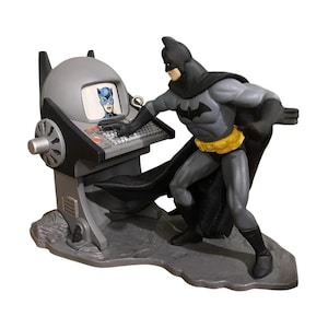 Può includere: Una statuetta di Batman in piedi davanti a una console grigia con tastiera nera e uno schermo che mostra un'immagine di Catwoman. Il personaggio indossa una tuta grigia e nera con una cintura gialla e un mantello nero su base grigia.