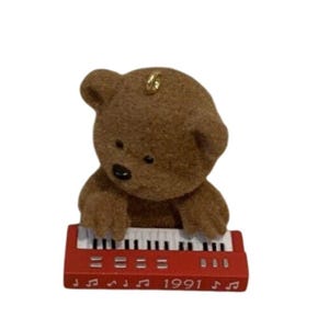 May include: A brown fuzzy bear figurine playing a red electronic keyboard. The keyboard features white and black keys, silver buttons, and the year 1991 printed in white along the front edge, accompanied by small white musical note symbols.