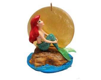 Puede incluir: Una figura decorativa de una sirena con largo cabello rojo y una cola verde brillante, sentada sobre una base de roca marrón rodeada de agua azul. Detrás de la sirena hay un gran disco circular de color amarillo translúcido.